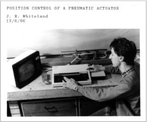 Picture of a university student siting beside a BBC Micro computer (and its small monitor) sitting on a concrete bench in front of a bit of equipment (a pneumatic cylinder, it's tubes and control lines)