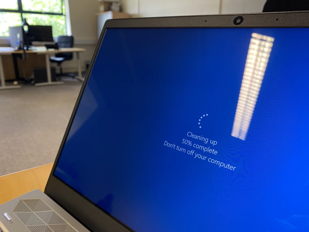 PC laptop displaying a blue screen saying "Cleaning up 50% complete Don't turn off your computer". It's sitting on a desk in an office, reflections of the office lighting are prominent on its screen.
