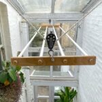 An airer hanging in a glassed-in space. it has wooden brackets and white metal rods and is hanging from ropes and pulleys.