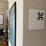 A square representation of a white keyboard key with the command symbol on it hanging on a wall in a room with other pictures, shelves, a desk and a computer
